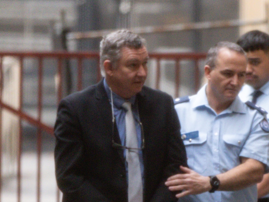 Greg Lynn wearing a suit and glasses around his neck being guided into court by a police officer.