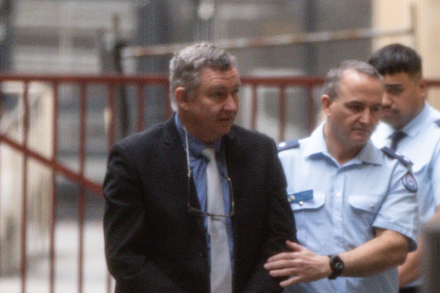 Greg Lynn wearing a suit and glasses around his neck being guided into court by a police officer.