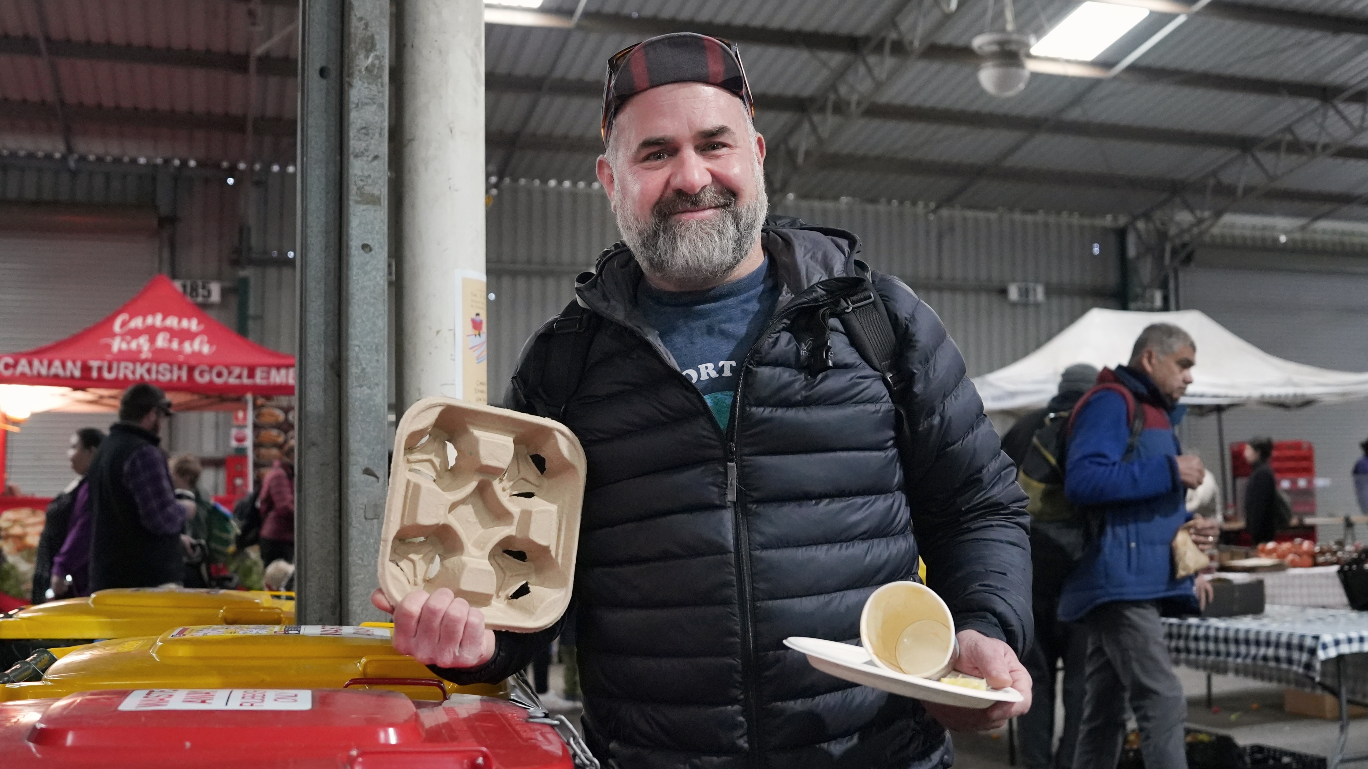 A man wearing a cap holding up compostable packaging in a warehouse surrounded by market stalls.