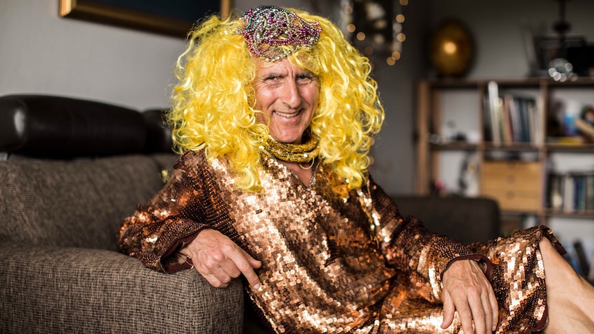 Kim Gotlieb on couch, wearing sequinned bronze dress and yellow-blonde wig.