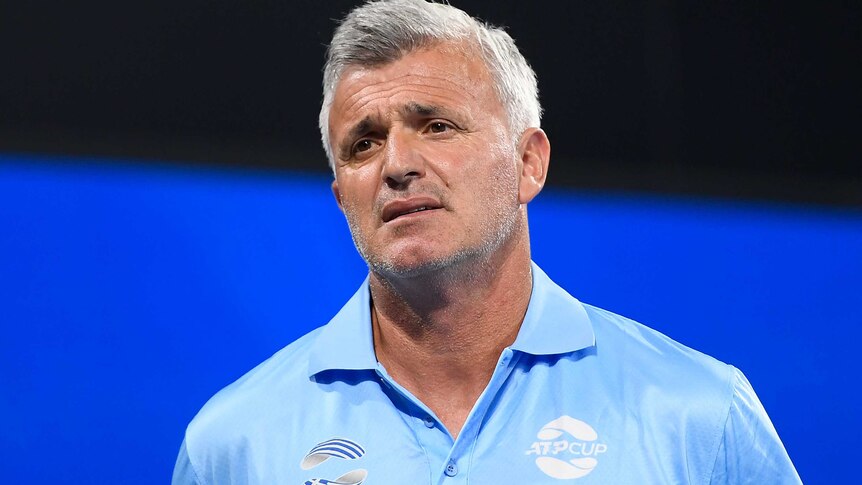 An older man with grey hair and wearing a light blue shirt has a pained expression on his face.