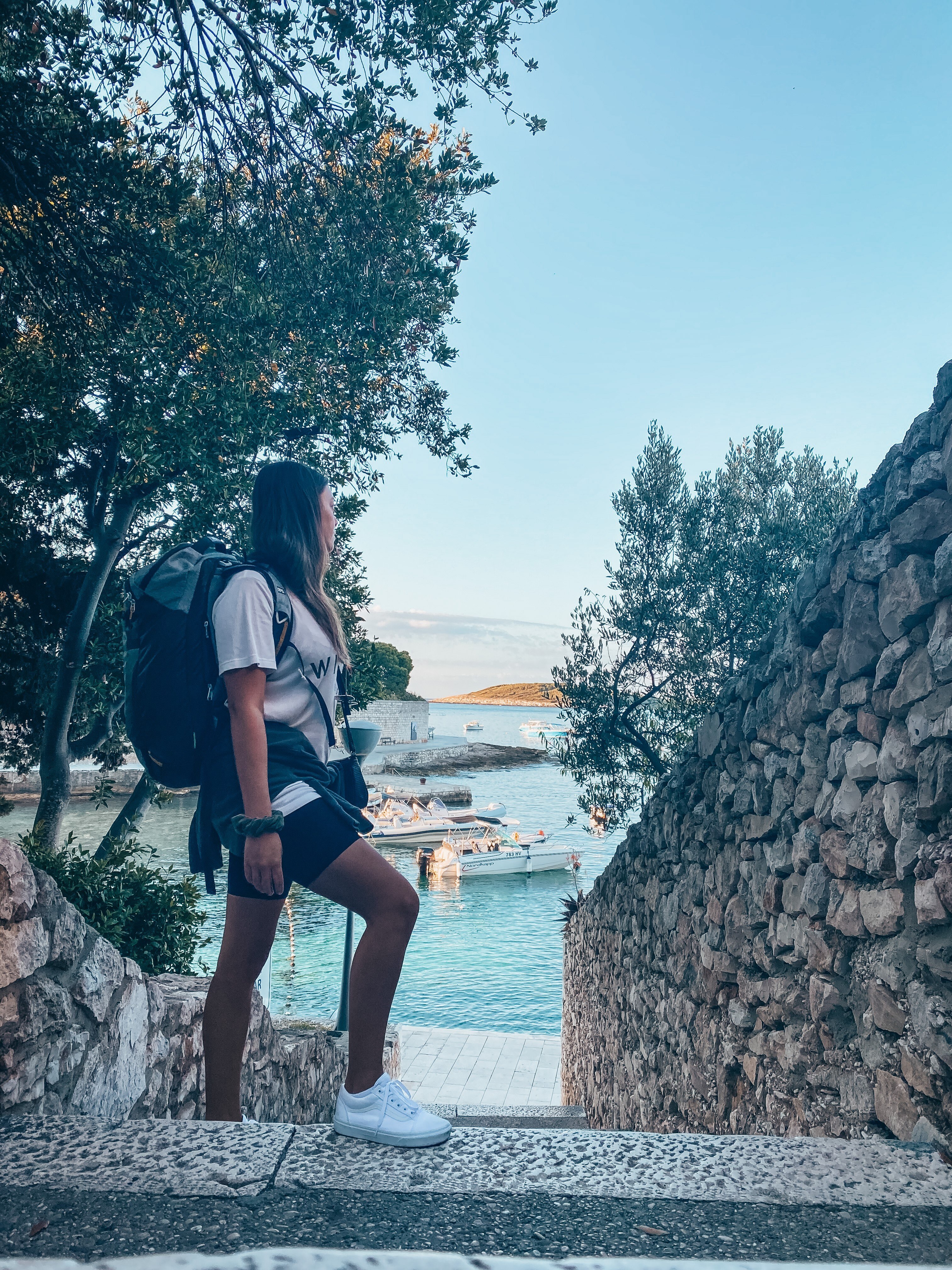 Kate wears a small carry-on backpack and looks out at a serene bay with boats while standing on stone steps..