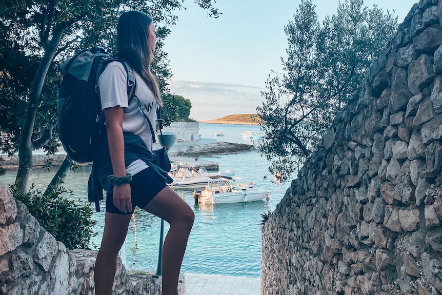 Kate wears a small carry-on backpack and looks out at a serene bay with boats while standing on stone steps..