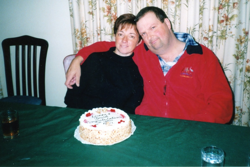 A woman and a man sitting together, embracing, in front of a birthday cake.