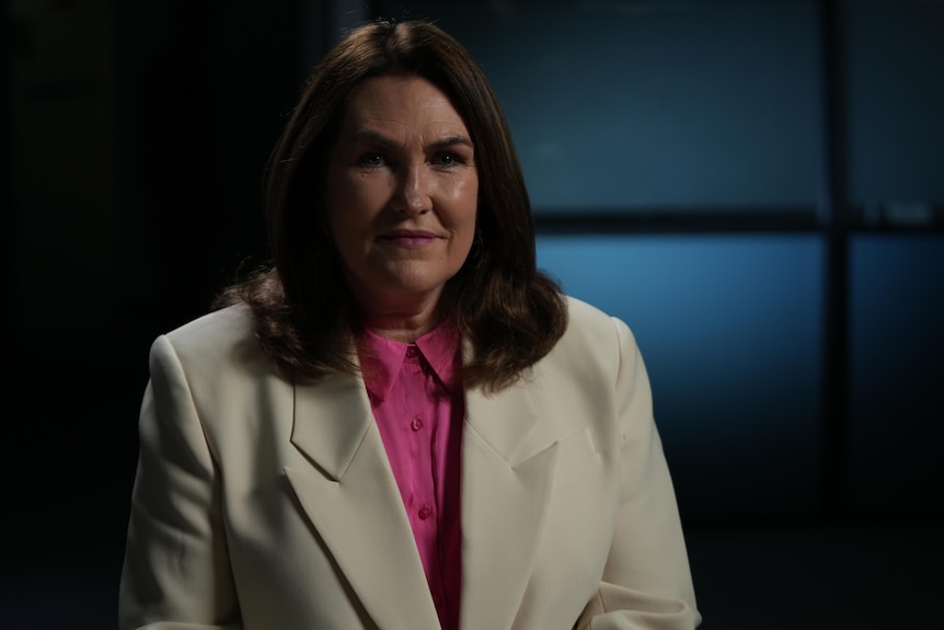 A woman with shoulder length brunette hair in a pink shirt with a white blazer