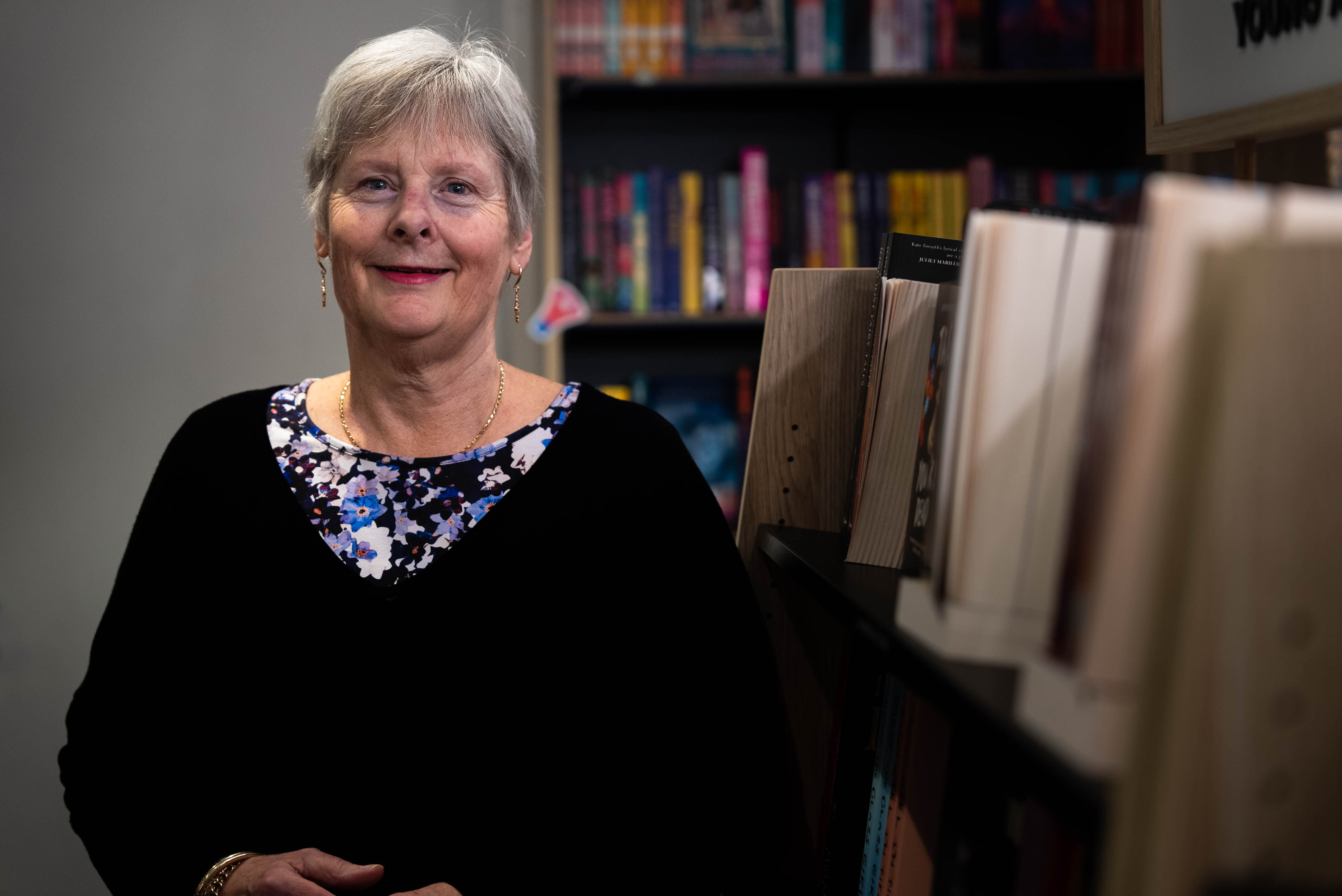 A woman with a black jumper smiles next to bookshelves.