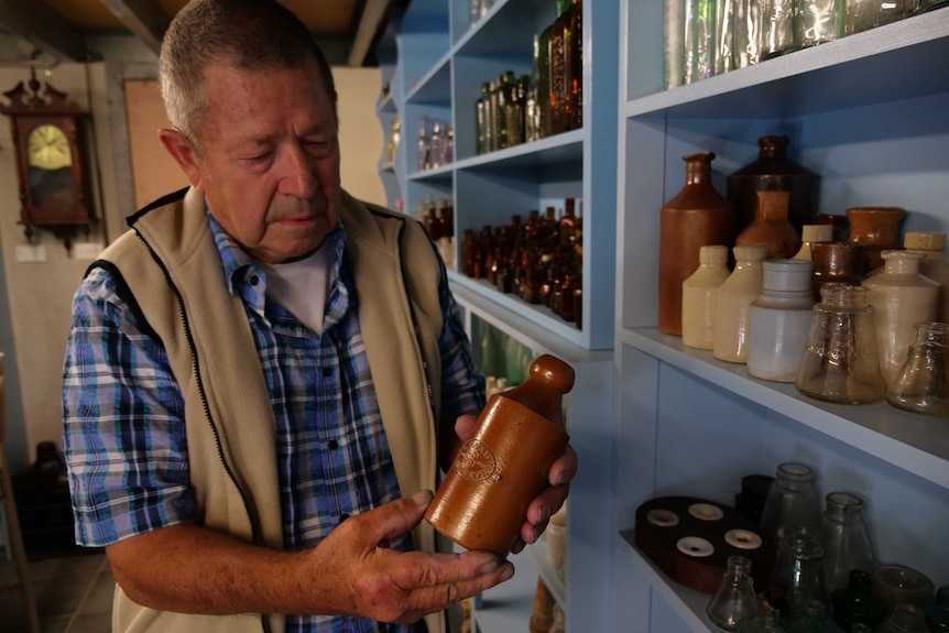 A man holding a clay bottle with shelves of glass bottles behind him.