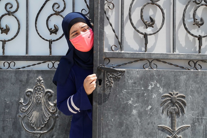 A woman wearing a hijab and a bright pink mask peaks out from behind an iron gate.