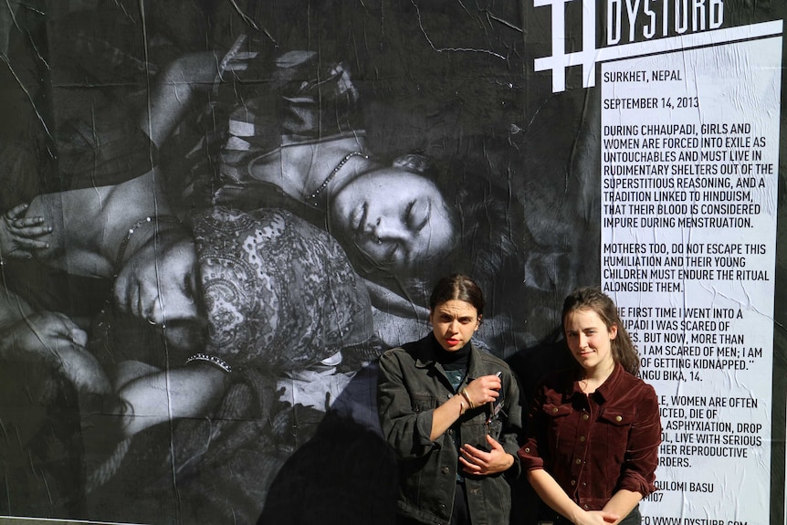 Dysturb's Madz Rehorec and Isabella Capezio stand in front of a large photograph of two Nepalese women and an infant child.