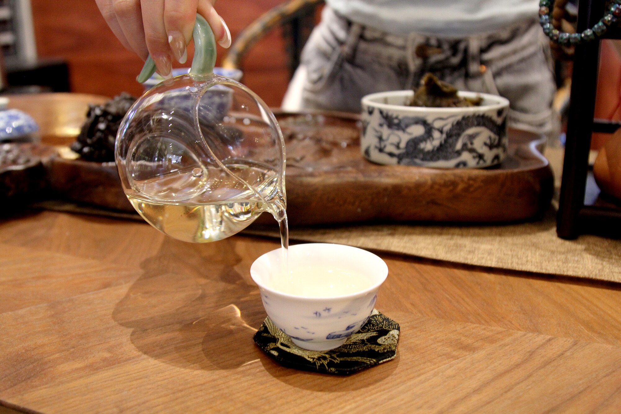 A person pours tea from a clear glass teapot into a small white tea cup.