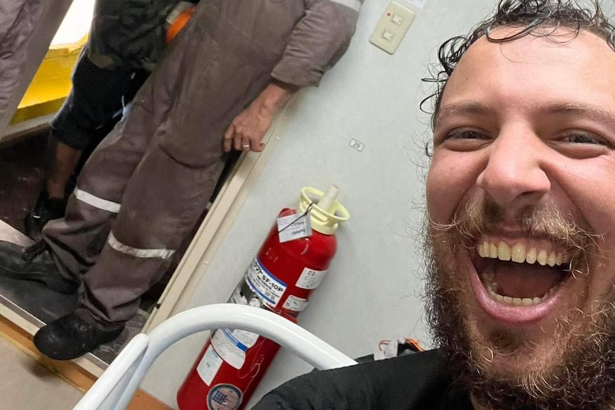 A smiling man sitting with a group of people in grey jumpsuits and white helmets in the background