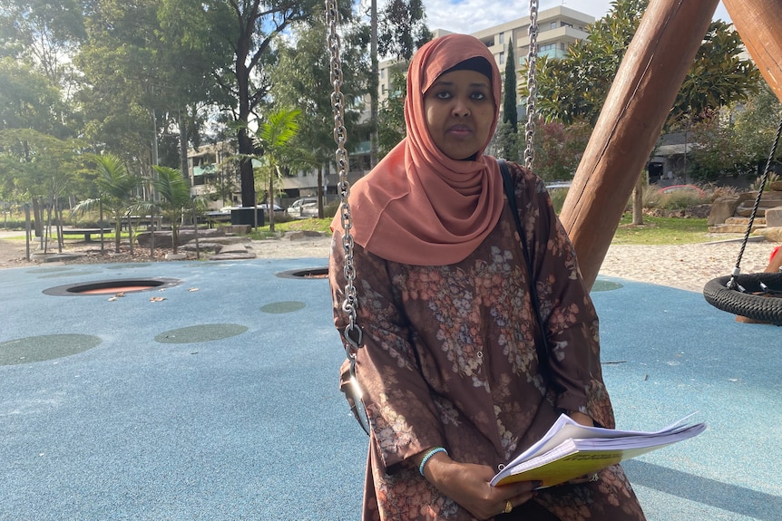 Ubah Scek, wearing a peach-coloured hijab, holds documents in her hands as she sits outside on a swing.