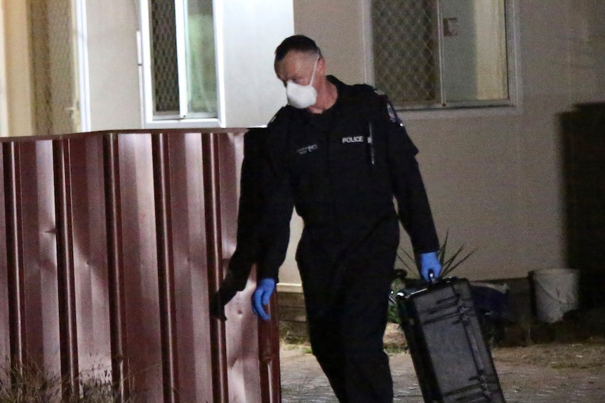 A police forensic officer walks out of Cleo Smith's house at night and around a fence carrying a black case.