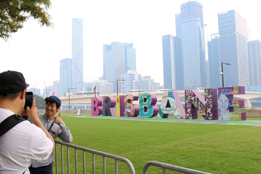 A man poses for a photo in front of the "Brisbane" sign in front of a hazy Brisbane skyline.