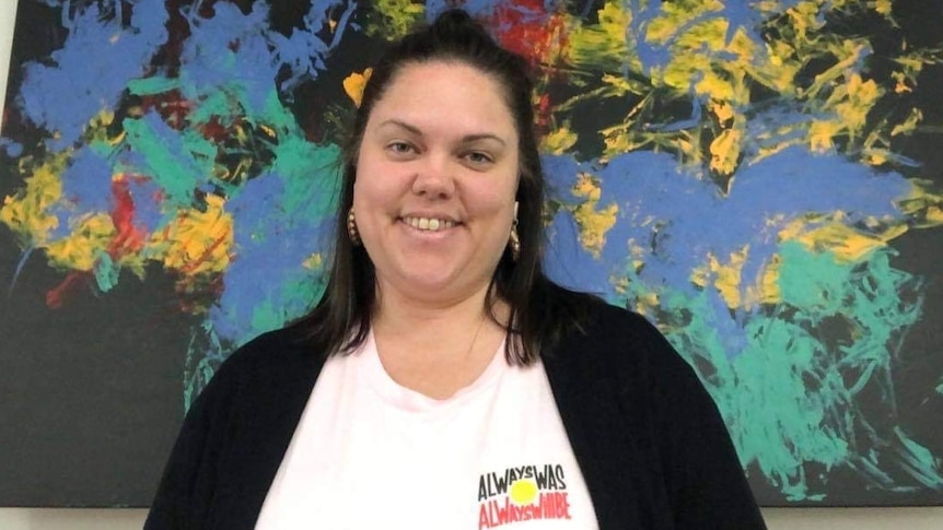 Rachael McPhail stands smiling at the camera in front of a large Indigenous art work behind her.