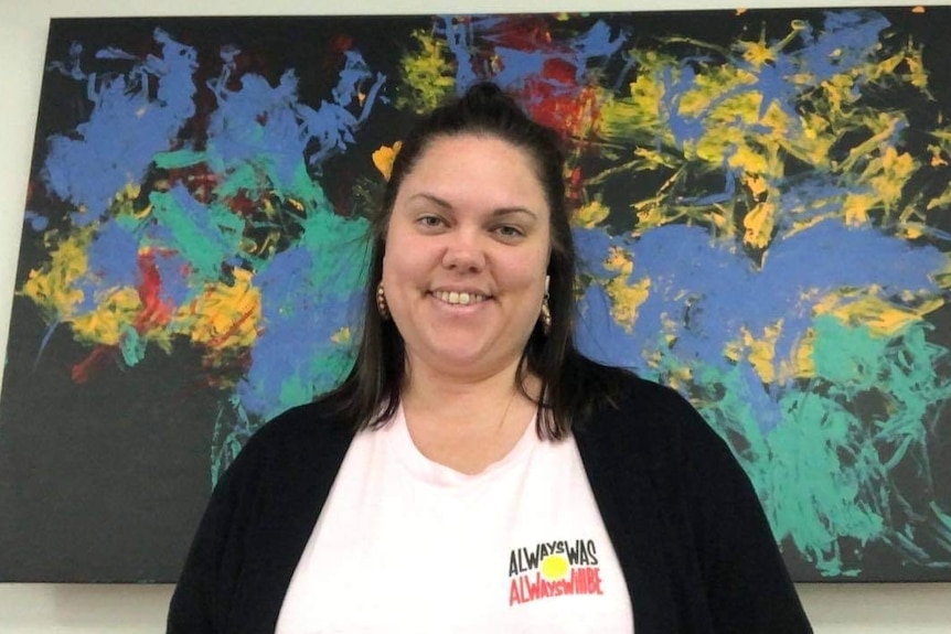 Rachael McPhail stands smiling at the camera in front of a large Indigenous art work behind her.