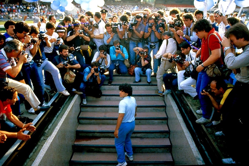A throng of cameramen surround Diego Maradona at the top of a set of stairs leading into a stadium.