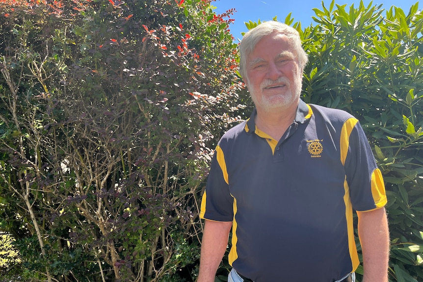 A man with grey hair and beard, wears a yellow and blue shirt, stands under a blue sky and in front of greenery.
