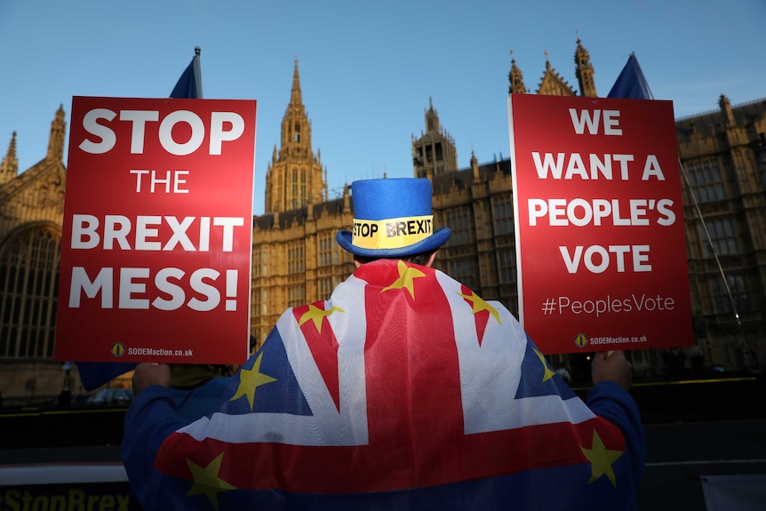 A man dressed in a top hat and flag draped over his shoulders holds two red placards opposing Brexit.