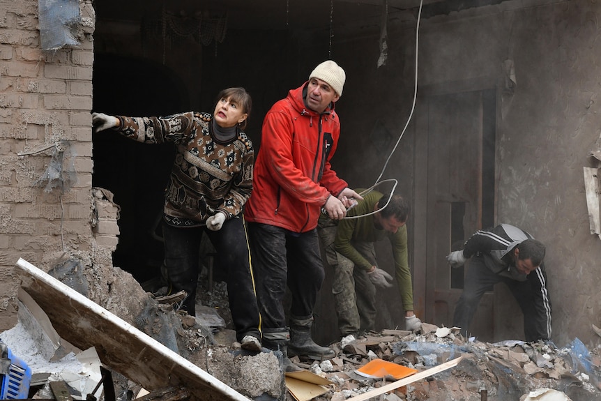 A man and woman peer out of a damaged home with debris surrounding them.