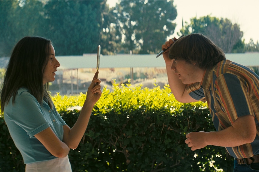 Both dressed in 70s-style clothing, a 20-something brunette woman holds up a mirror for a teenage boy to comb his hair