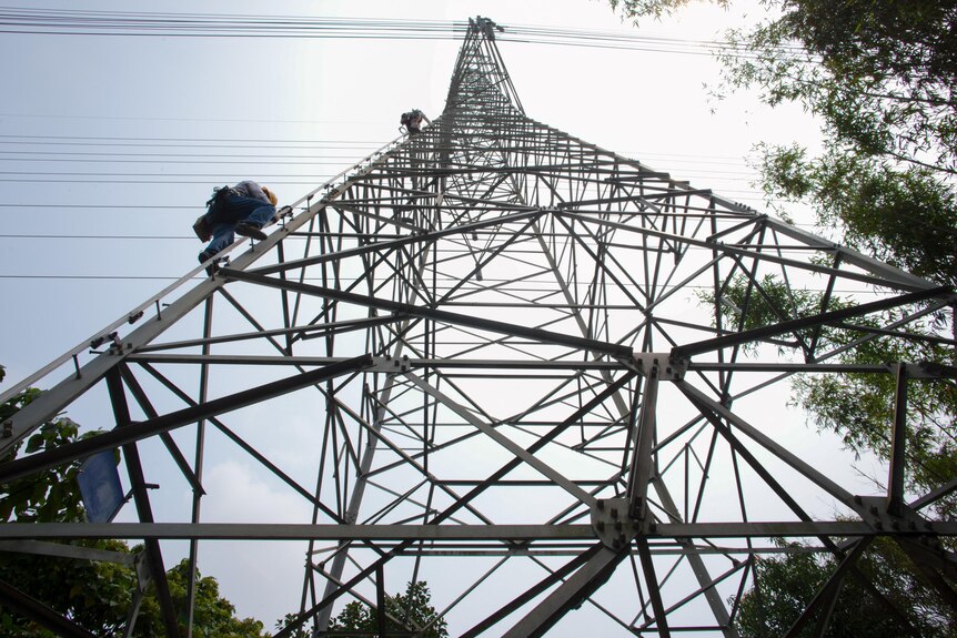 Workers climb a huge electric tower that rises far into a clear sky.