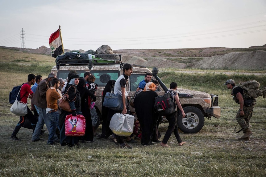 Families holding bags walk beside a battered armoured vehicle flying the Iraqi flag