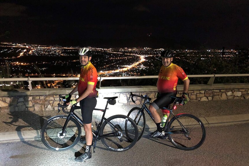 Two cyclists wearing lycra stand next to their bikes on top of Townsville's monolith Castle Hill, it is pre-dawn.
