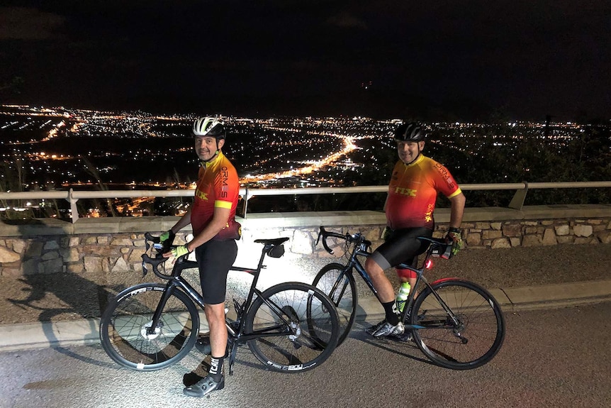 Two cyclists wearing lycra stand next to their bikes on top of Townsville's monolith Castle Hill, it is pre-dawn.