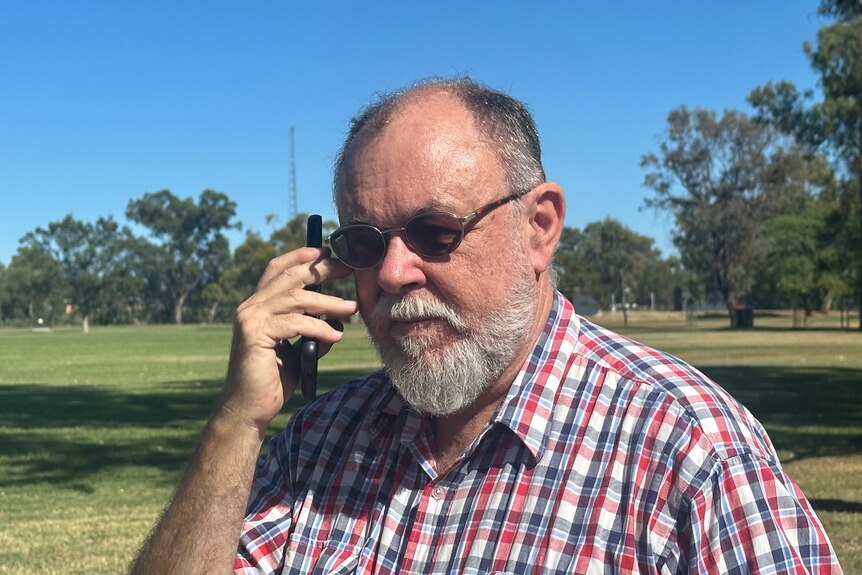 A close up of a man in his 60s with a beard and glasses, making a phone call outside near a grass field.