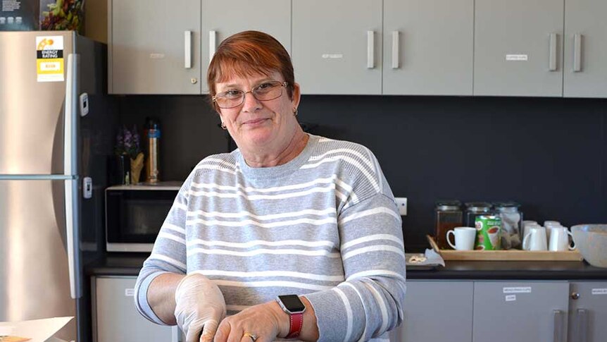 A woman with short coppery hair and glasses in a kitchen cuts up an uncooked sausage roll
