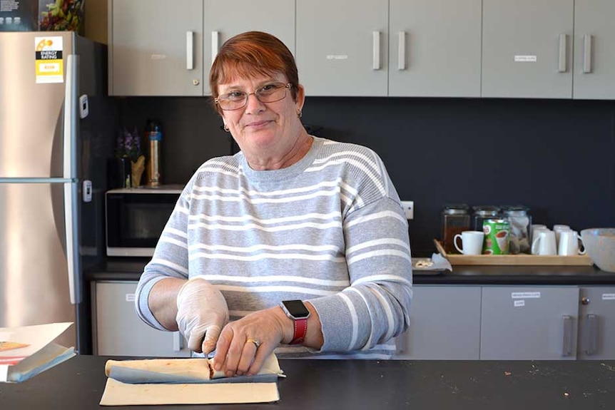 A woman with short coppery hair and glasses in a kitchen cuts up an uncooked sausage roll.