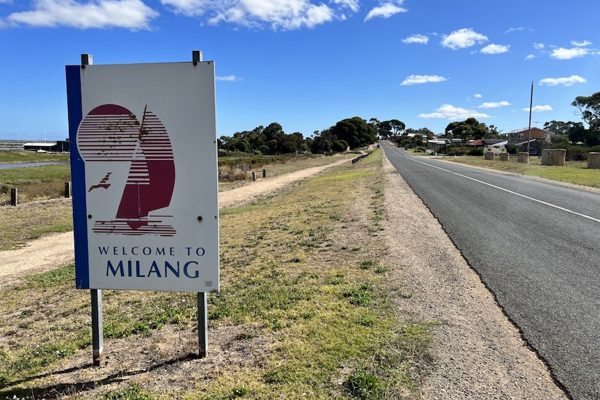 A community welcome sign reading 'Milang', next to a road running alongside the shore of a lake