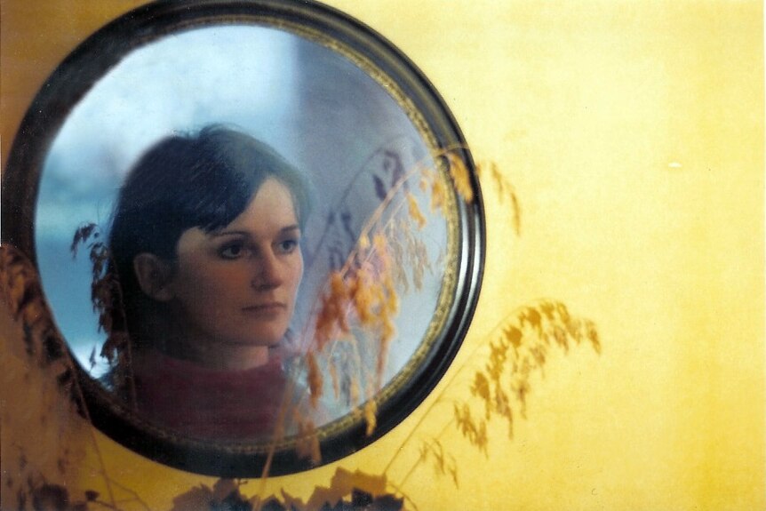 A young Amanda Feilding looks into a circle mirror hung on a yellow wall.