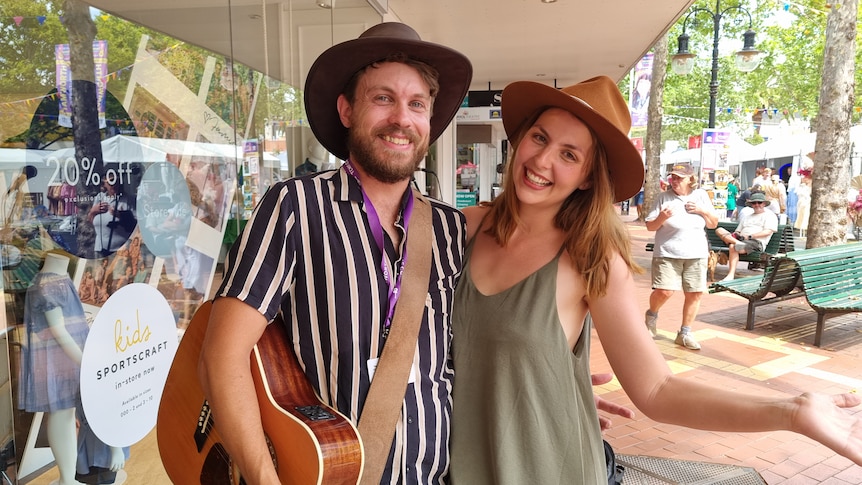 Two people standing on a footpath wearing hats, one holding a guitar.