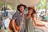 Two people standing on a footpath wearing hats, one holding a guitar.