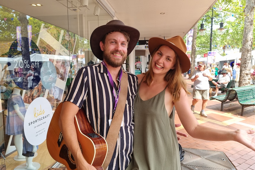 Two people standing on a footpath wearing hats, one holding a guitar.