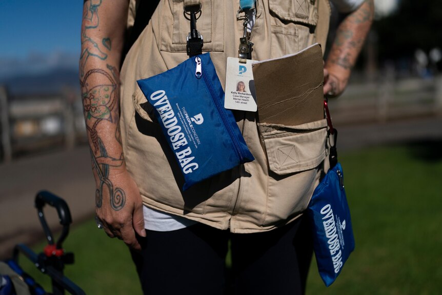 A woman's torso, her arms tattooed, with two blue 'overdose bags' hanging off the pockets of her vest. 