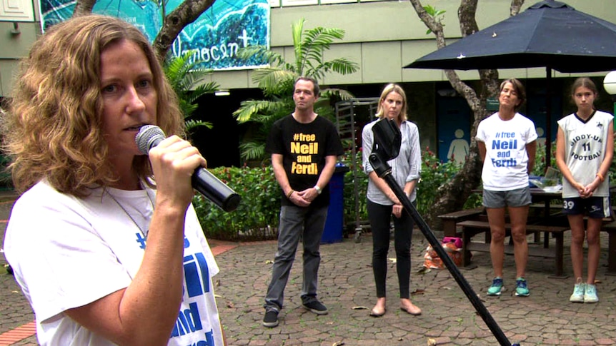 Woman with microphone stands in the foreground with three people listening behind her.