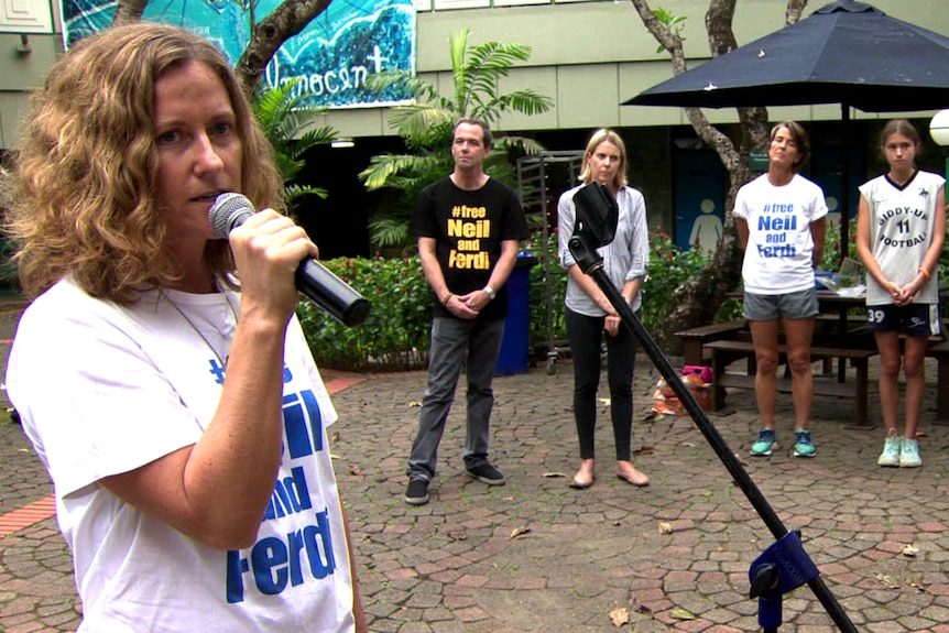 Woman with microphone stands in the foreground with three people listening behind her.