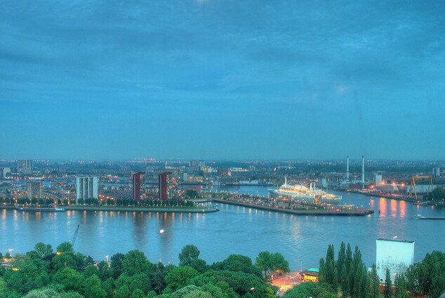 A misty early-evening aerial image of a port with a bluish tinge to the clouds and pink lights shining on the water
