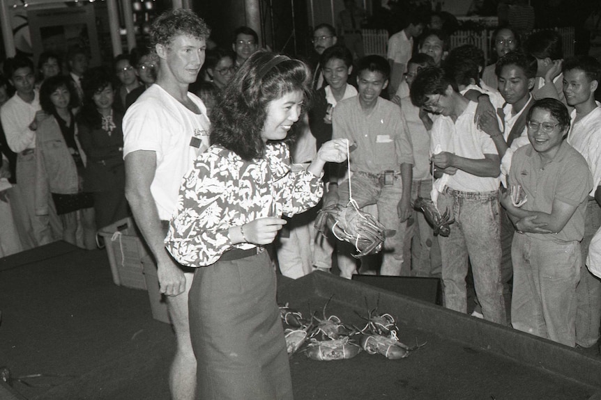 A woman in business attire holding a tied-up mudcrab before a wowed audience. Billy Lowey behind.