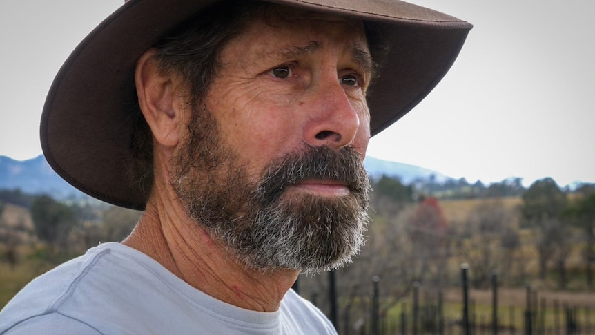 A man with a salt-and-pepper beard wearing a broad-brimmed hat stares off into the distance.
