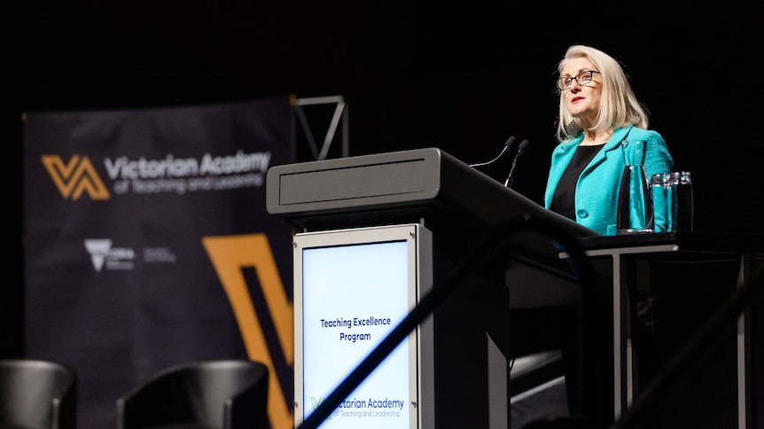 A woman wearing a turquoise-coloured suit jacket and glasses stands at a lectern as she delivers a speech.