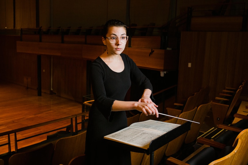 Nicky Gluch wearing black and conducting in dimly lit music hall.