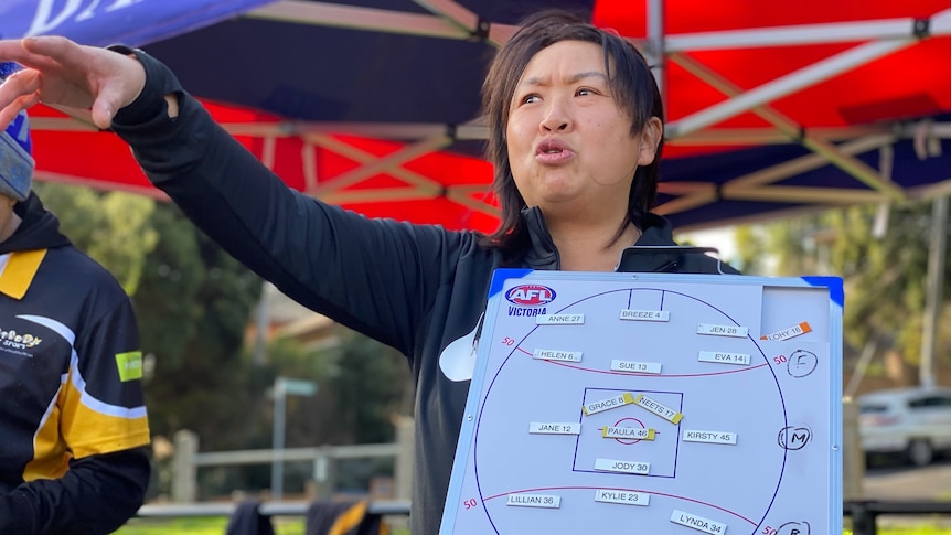 Sarah Loh holds a whiteboard with a diagram of an Australian Rules playing field and players' names.