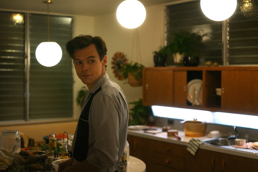 Brunette white man wearing grey shirt looks over shoulder in a 50s-styled kitchen interior.