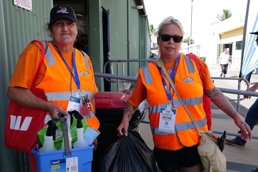 Cassandra and Natalie in orange high vis vests, holding a rubbish bag and cleaning tools.