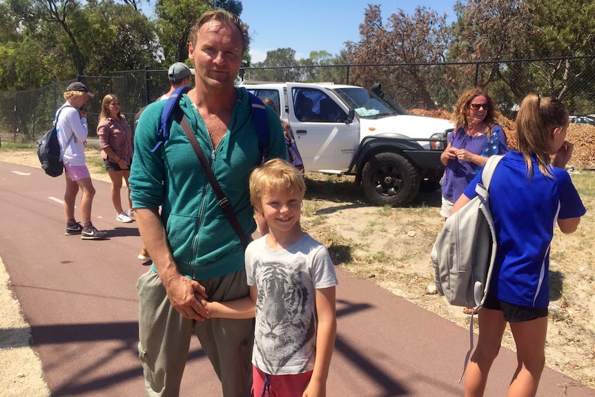 A man stands with his son on the footpath outside a school as other parents and children linger neaby.