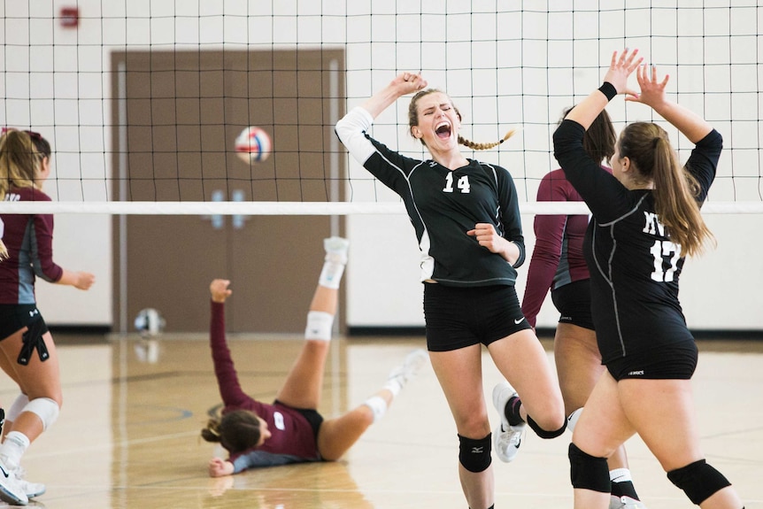 A group of young women jump in celebration near a volleyball net.